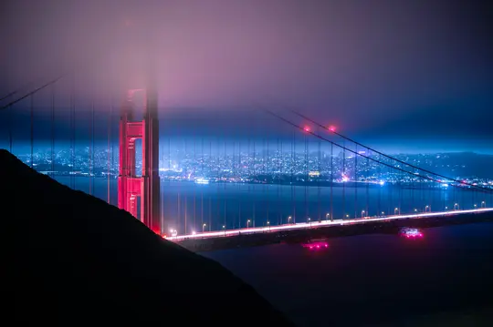 Una foto de larga exposición del Puente Golden Gate por la noche, con una torre roja parcialmente oculta por la niebla. Las