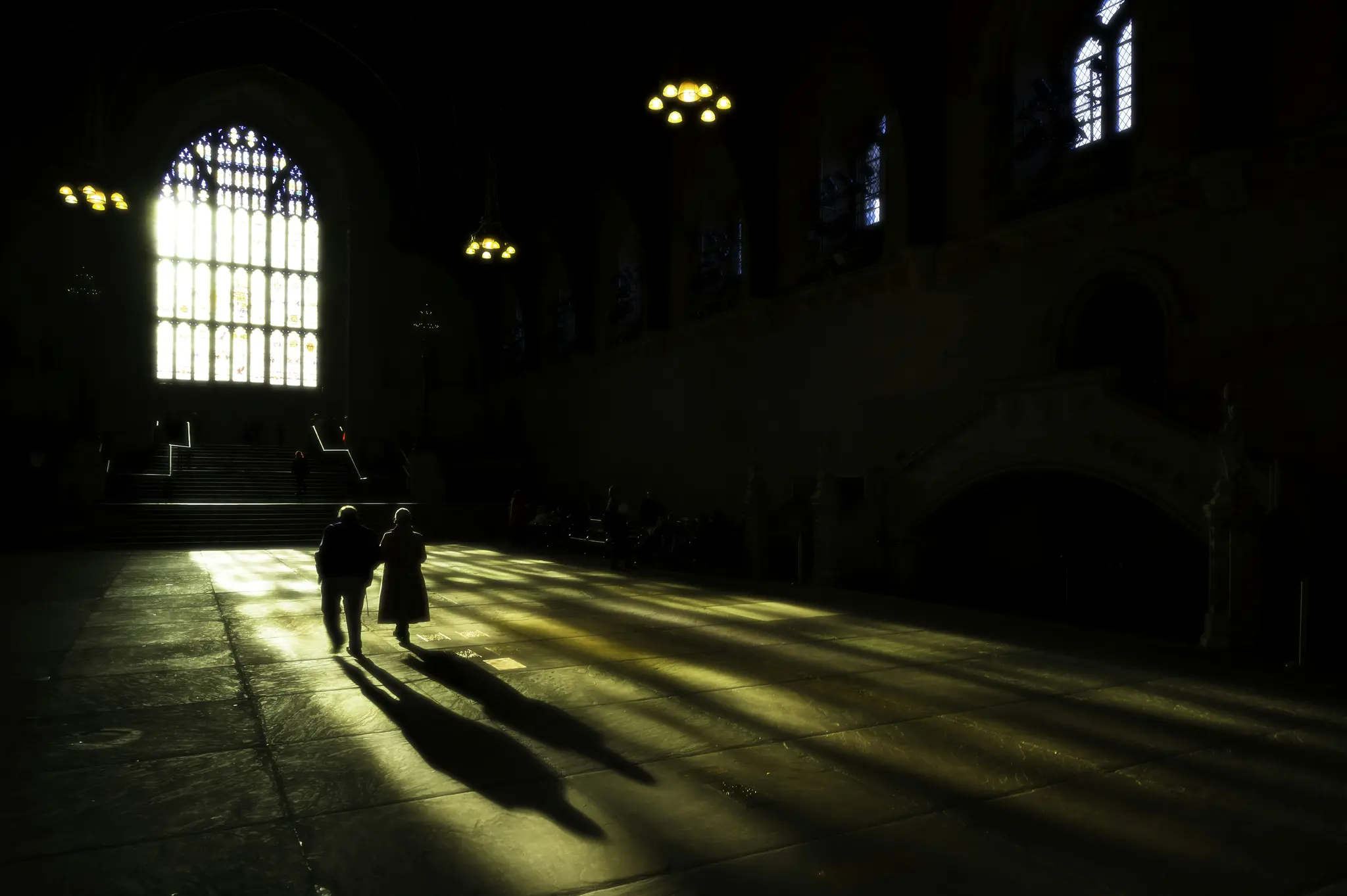 Twee silhouetten lopen over een uitgestrekte, donkere hal, verlicht door fel licht dat door een groot glas-in-loodraam