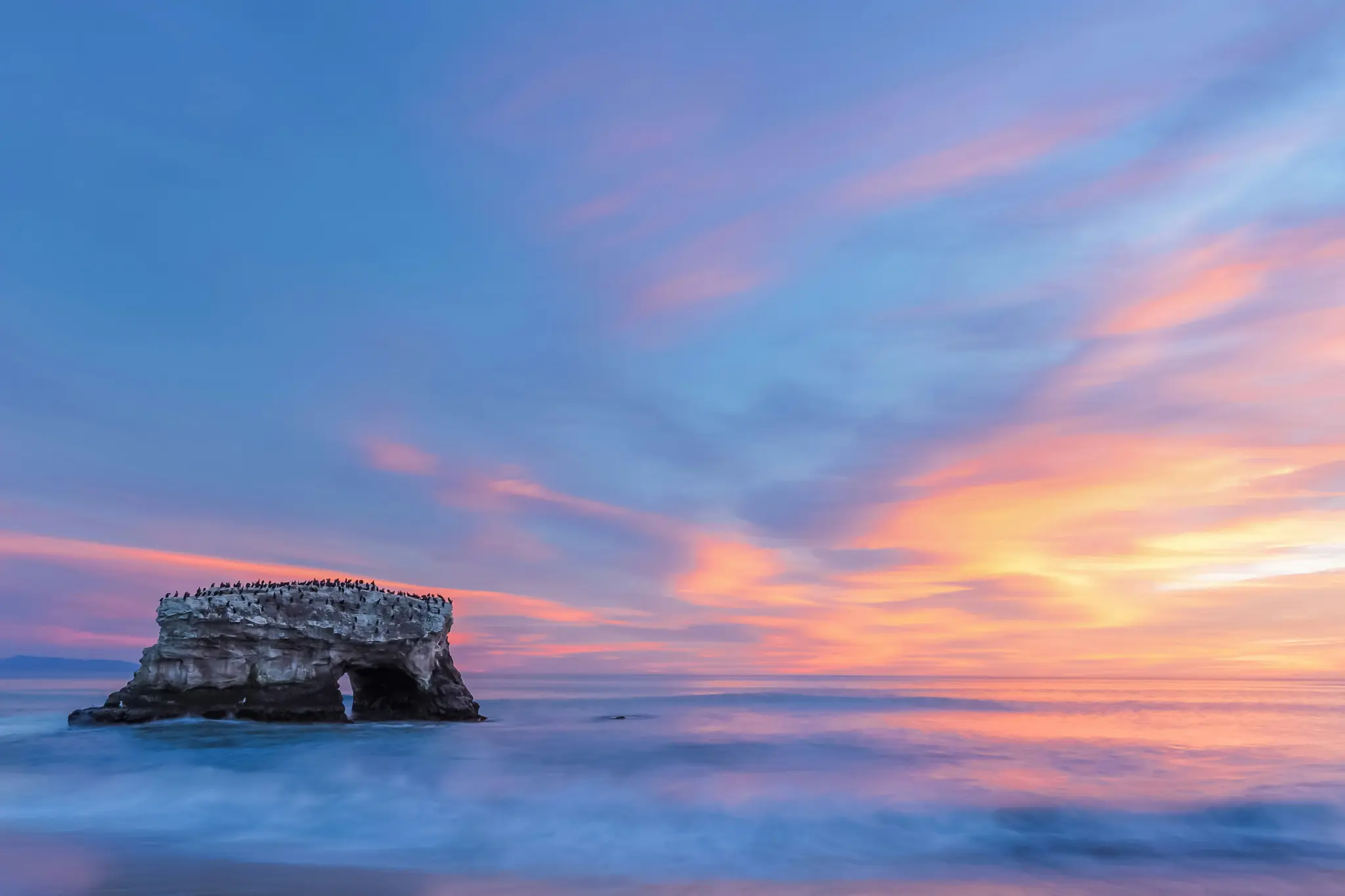 Een brede landschapsfoto van een natuurlijke rotsboog in de oceaan bij zonsondergang. De lucht is levendig met blauwe, roze