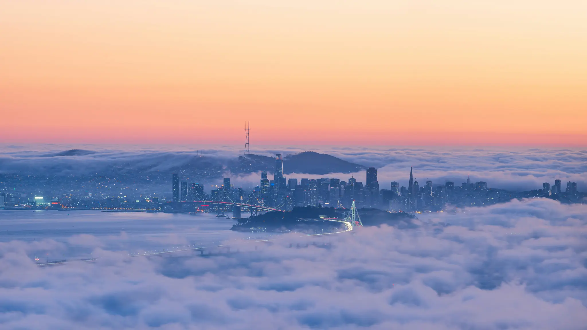 Et luftfotografi af et landskab viser en bys skyline, der dukker op af et tykt lag hvid tåge under en himmel, der skifter