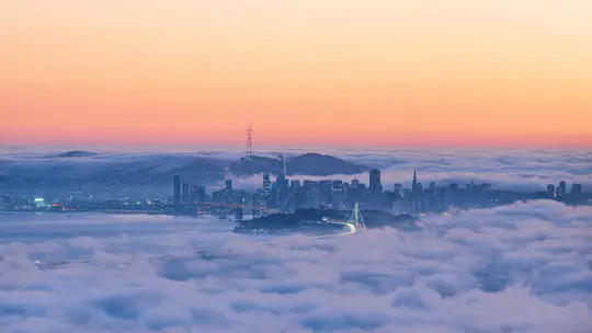 Letecká krajinářská fotografie ukazuje panorama města vynořující se z husté vrstvy bílé mlhy pod oblohou přecházející z...