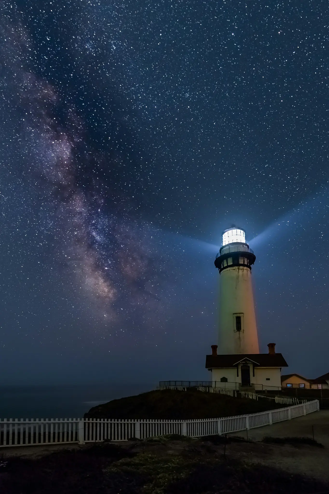 Un alto faro bianco si erge su una scogliera buia sotto un vasto cielo notturno pieno di stelle con la Via Lattea che si