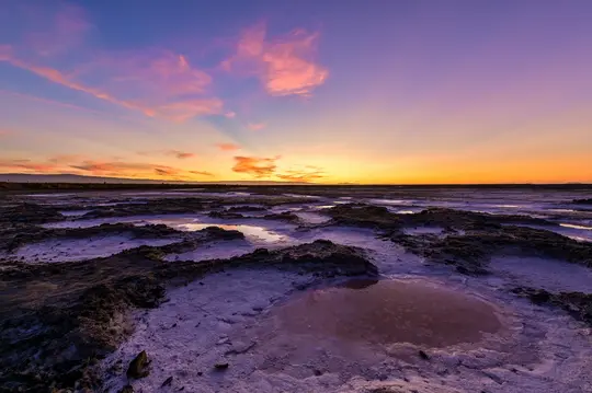O fotografie largă de peisaj cu saline la apus. Cerul este un gradient vibrant de violet, roz, portocaliu și albastru.