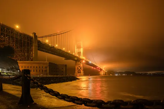 A Golden Gate híd drámai éjszakai felvétele, amelyet sűrű, narancssárga köd takar, a vízen tükröződő fényekkel.