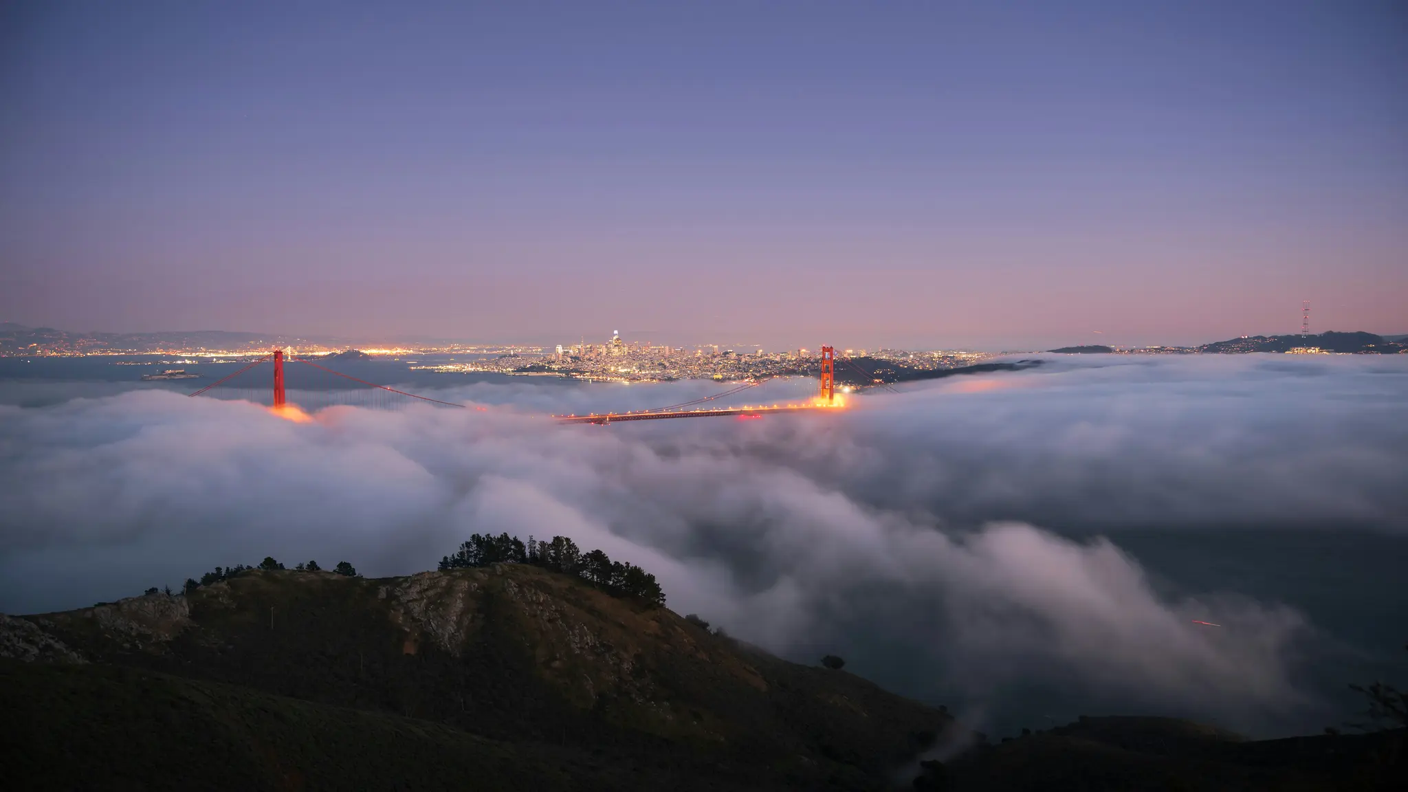 Een luchtfoto van de Golden Gate Bridge en de skyline van San Francisco bij schemering, gedeeltelijk bedekt door dichte mist.