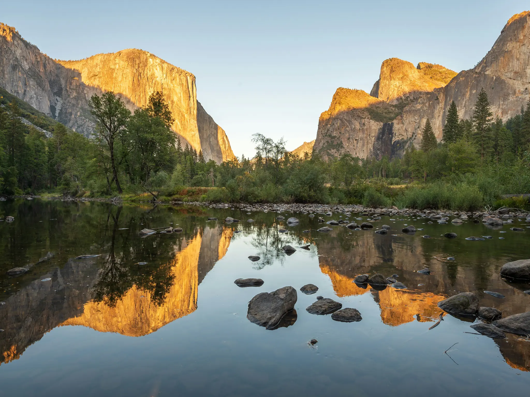 Široká krajinárska fotografia Doliny Yosemite. Pokojná rieka v popredí odráža slnkom zaliate žulové útesy a zelené stromy.