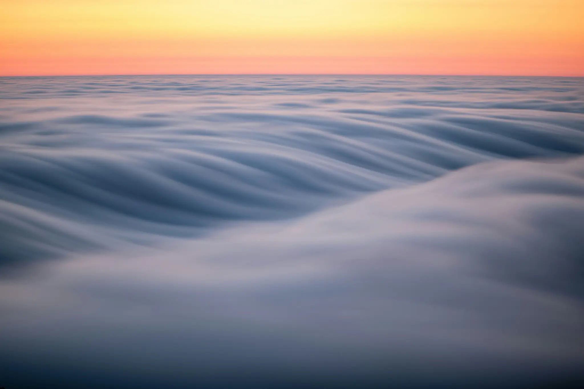 A long exposure shot of a vast expanse of soft, wavy clouds under a sky transitioning from orange to yellow.
