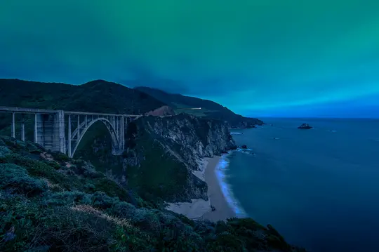 Een lange belichting van de Bixby Bridge die een diepe kloof overspant langs de Pacifische kust. De lucht is turkoois, de