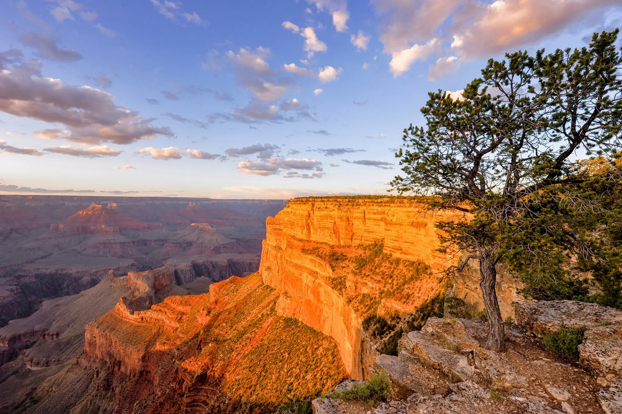 Széles tájfotó a Grand Canyonról naplementekor, hatalmas narancssárga és vörös sziklaformációkat mutatva kék ég alatt