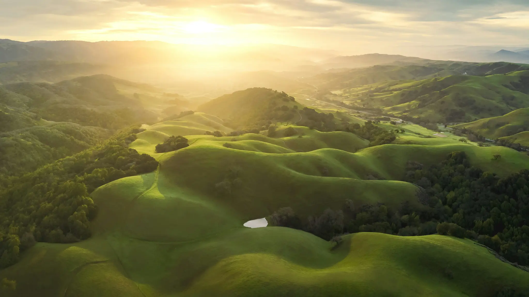 Een luchtfoto toont uitgestrekte, glooiende groene heuvels en valleien onder een heldere, warme zonsonderganglucht, die lange