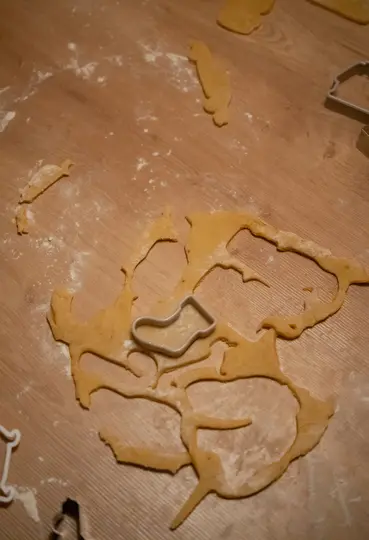 An overhead close-up shows cookie dough and cookie cutters on a wooden surface. Flour dusts the wood and dough.