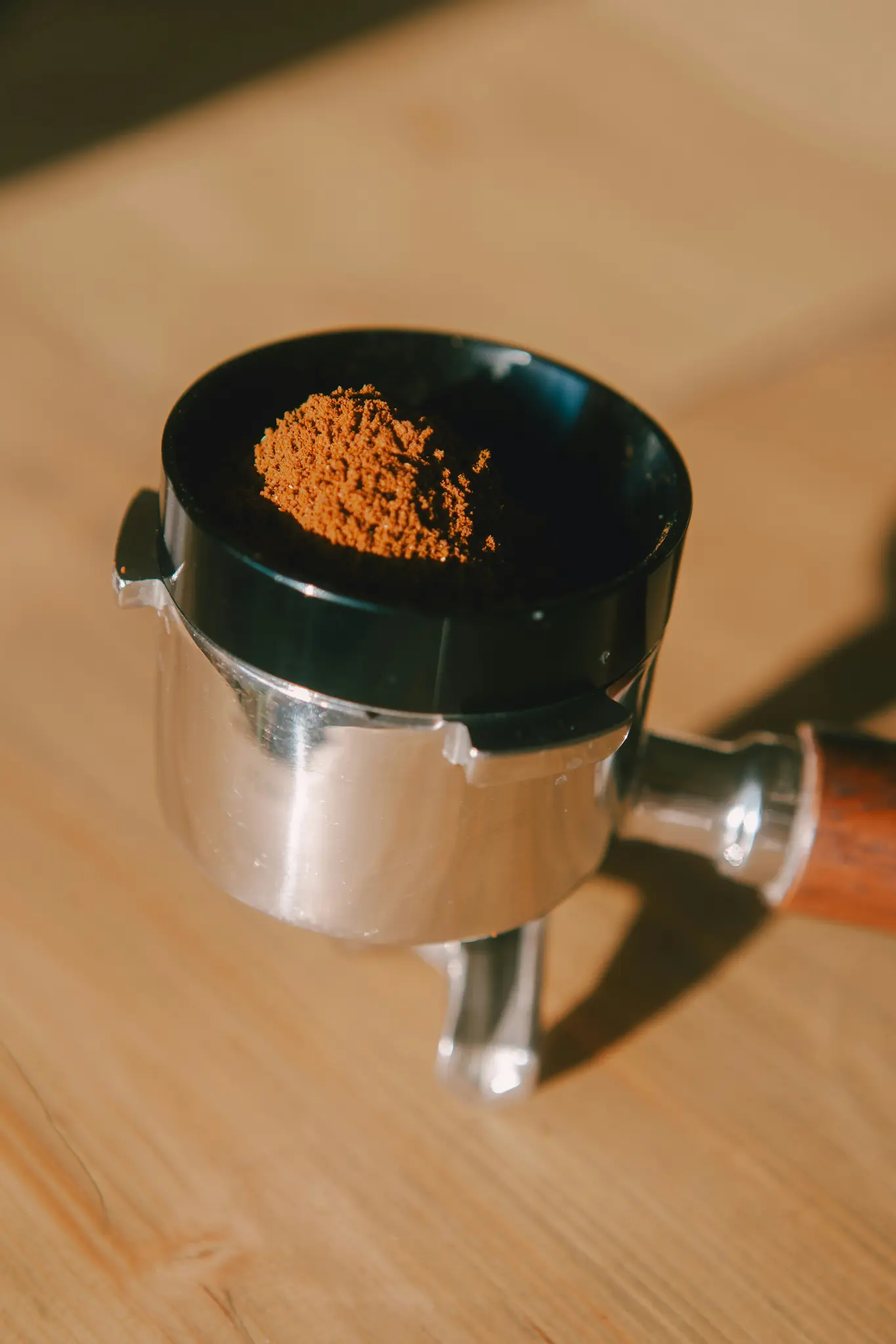 Detail stříbrného portafiltru s černým dávkovacím trychtýřem naplněným jemně mletou hnědou kávou, položený na světlém…