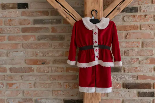 A red Santa Claus costume, complete with white fluffy trim and a black belt, hangs on a black hanger from a wooden beam.