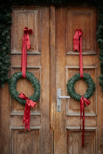 Two green Christmas wreaths with long red satin ribbons hang on a rustic wooden double door. A silver door handle is visible.