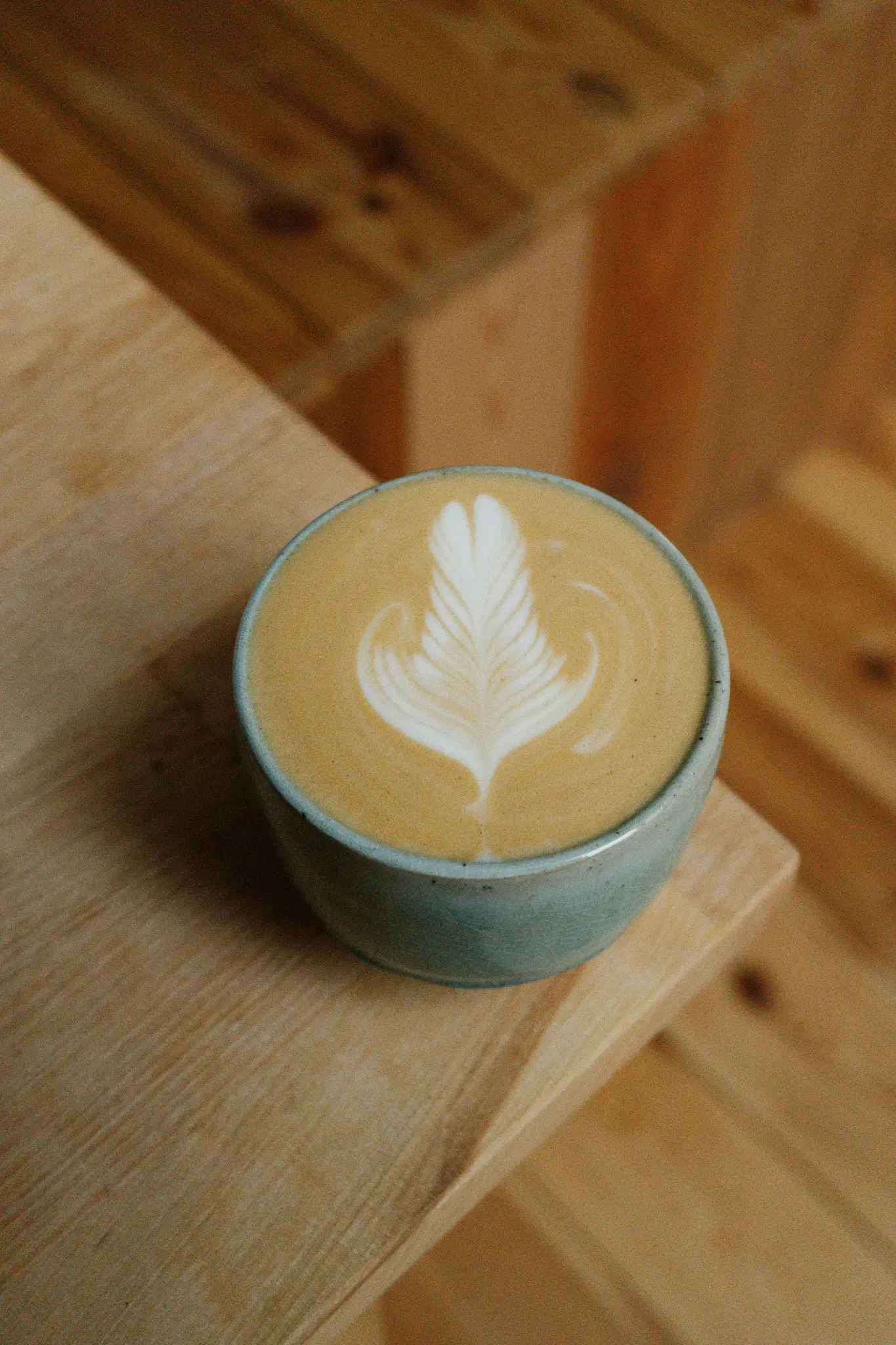 En ljusblå keramisk mugg med latte och intrikat vit fjäderformad lattekonst sitter på en ljusbrun träyta.
