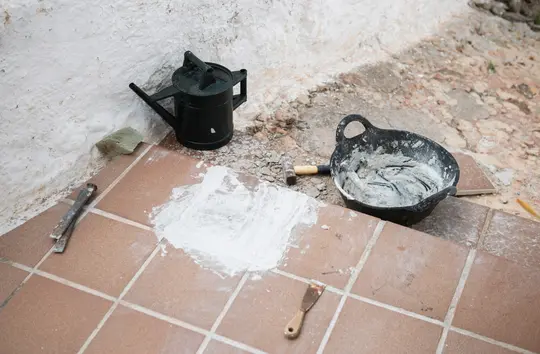 A black watering can, a black bucket filled with grey mortar, a hammer, two chisels, and a small spatula are on a brown…