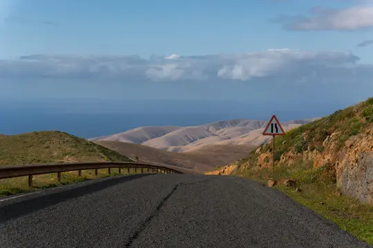 Una strada asfaltata scura serpeggia verso il basso attraverso colline secche, verdi e marroni verso un oceano blu sotto un