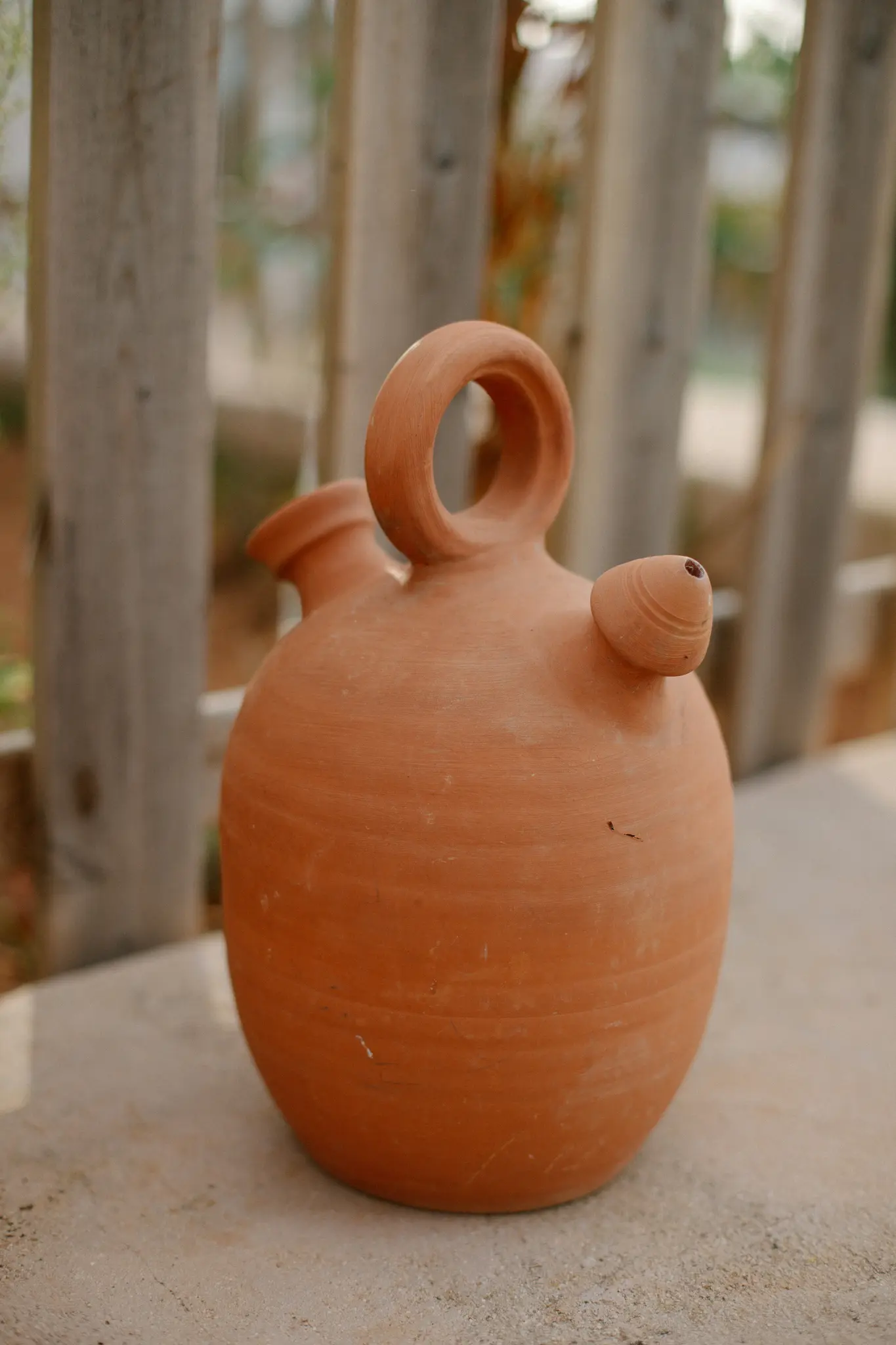 Et traditionelt terracotta botijo, et spansk drikkebeholder, står på en lys stenoverflade med en sløret træ...