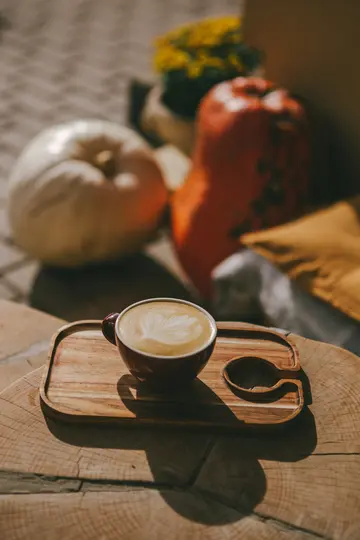 Una tazza di latte rosso scuro con un disegno a foglia nella schiuma si trova su un vassoio di legno marrone chiaro. Zucca