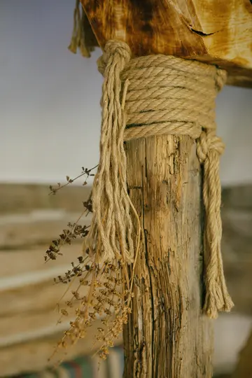 A close-up shot of a weathered wooden post wrapped with thick, natural-colored rope. Dried herbs hang from the rope.