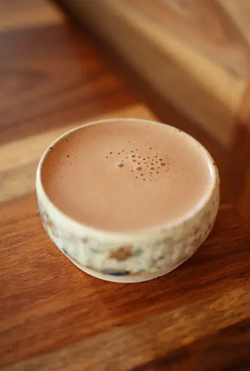 A light brown beverage with small bubbles on its surface fills a textured, light-colored ceramic bowl on a wooden table.