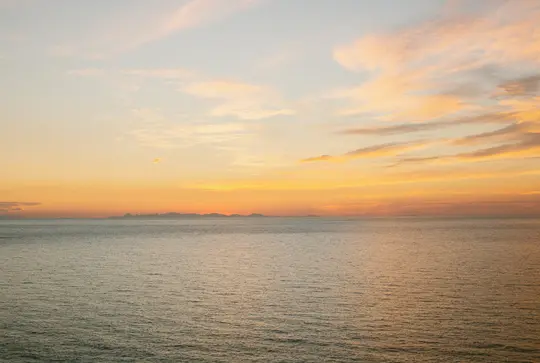 Un'ampia foto paesaggistica di un oceano calmo sotto un cielo al tramonto con sfumature arancioni, gialle e azzurre. Una