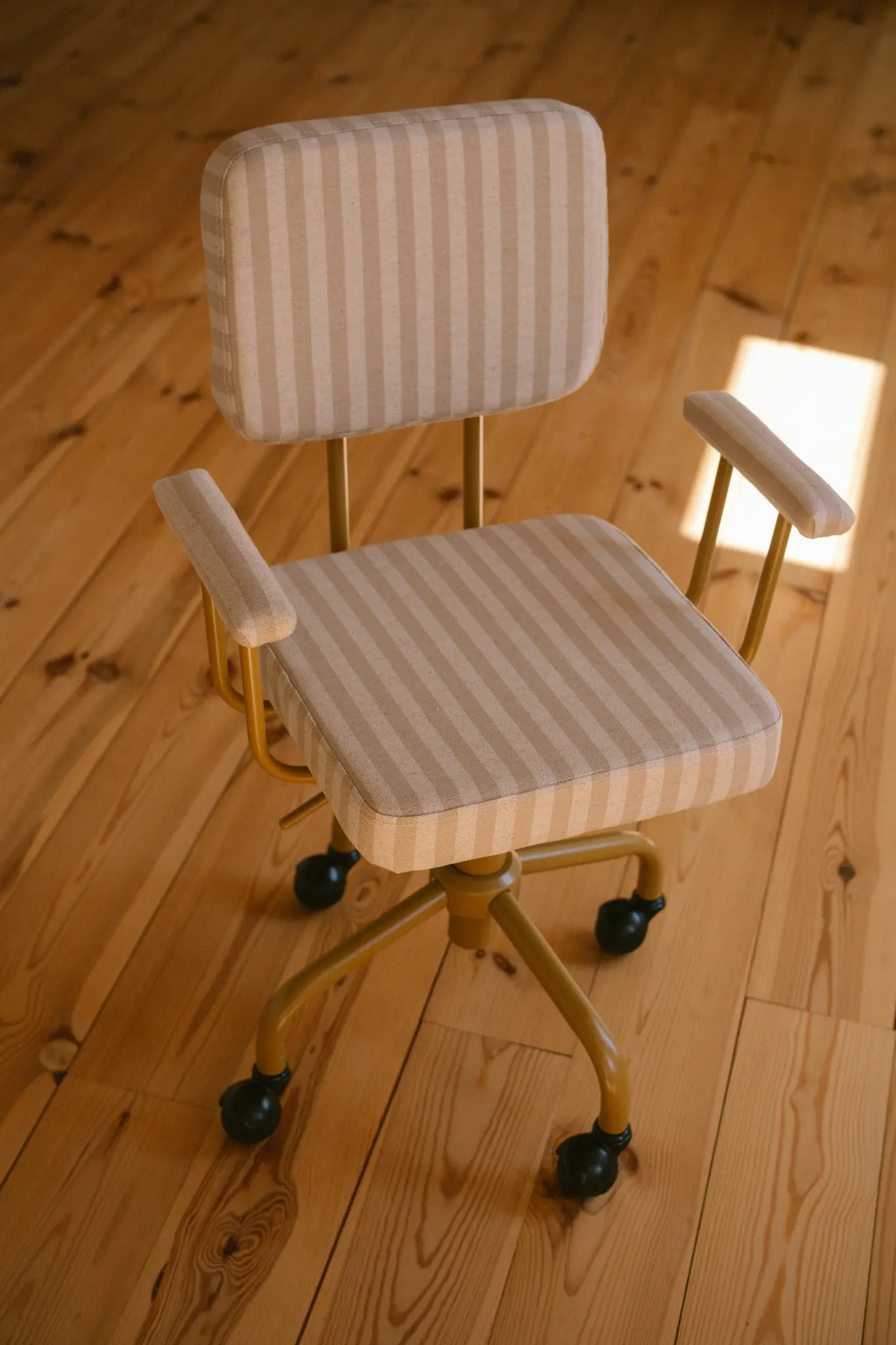 Een mosterdgele bureaustoel met beige en wit gestreepte bekleding staat op een lichte houten vloer, verlicht door een vlek