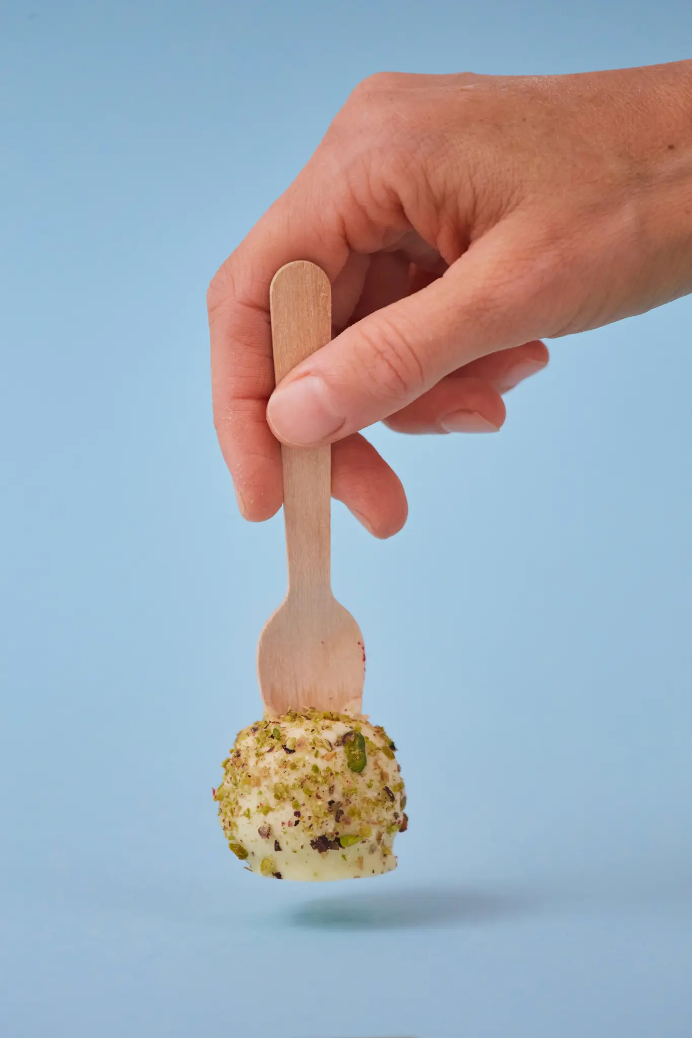 Eine Hand hält einen kleinen Holzlöffel mit einem weißen, runden Dessert, das mit grünen Pistazienstücken bedeckt ist, vor