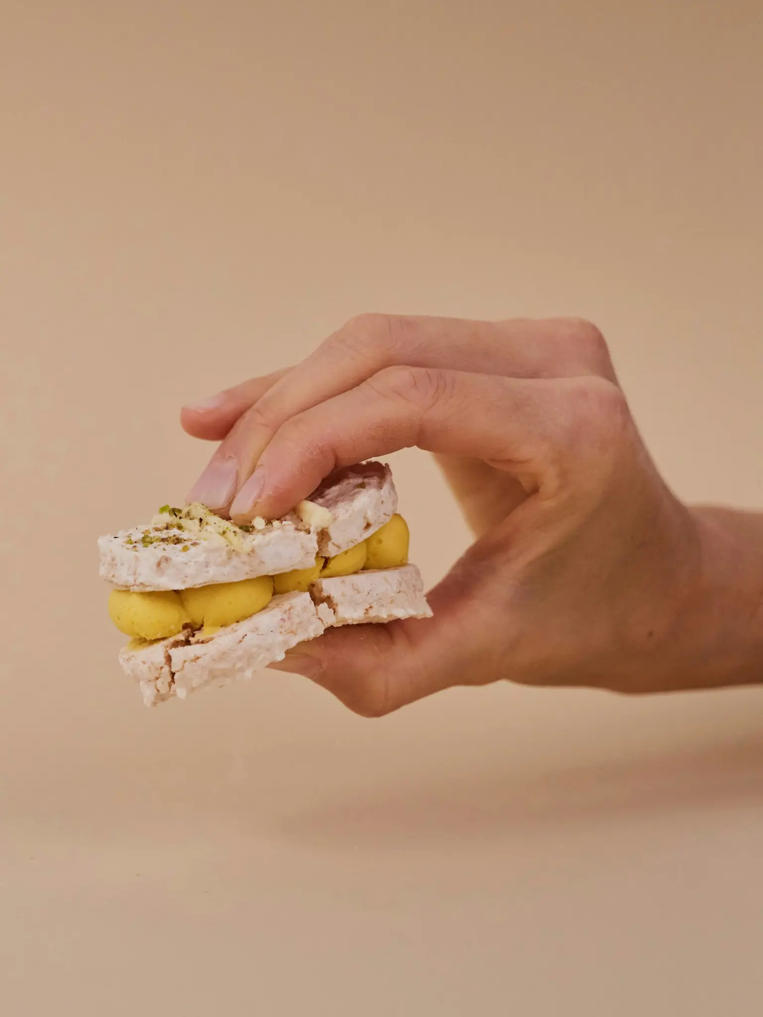 Una mano tiene un biscotto sandwich alla meringa con ripieno di crema giallo brillante e codette verdi su uno sfondo beige