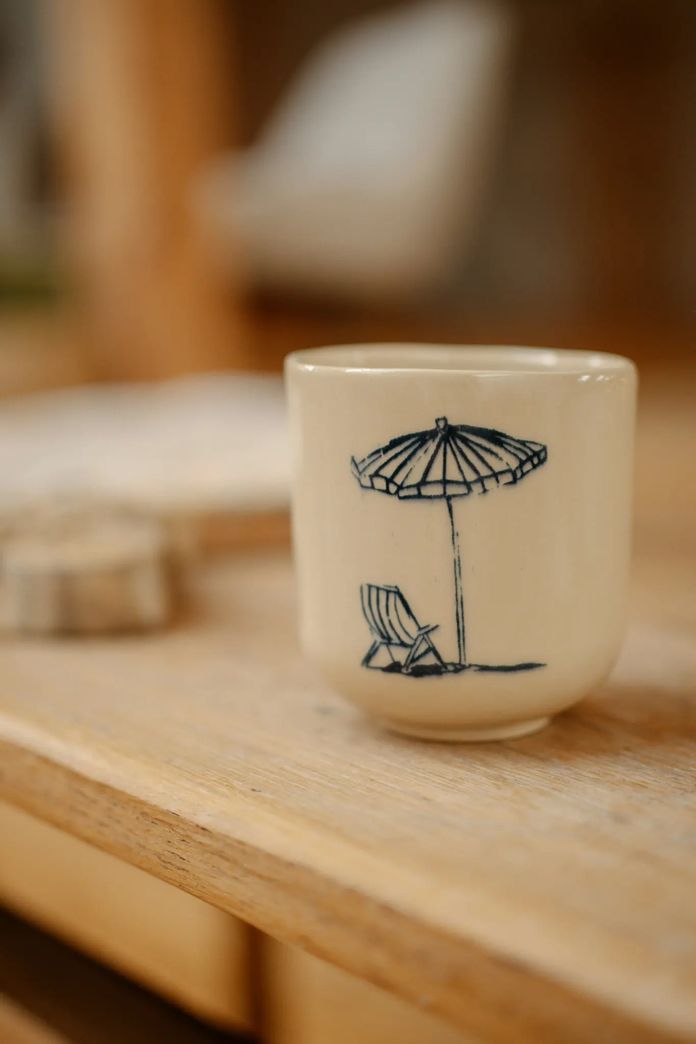 Une tasse en céramique de couleur crème repose sur une table en bois, ornée d'une illustration bleu foncé d'un parasol de