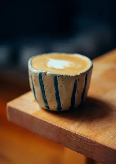A close-up shot of a ceramic cup with blue vertical stripes, holding a latte with a heart-shaped foam design, resting on a…