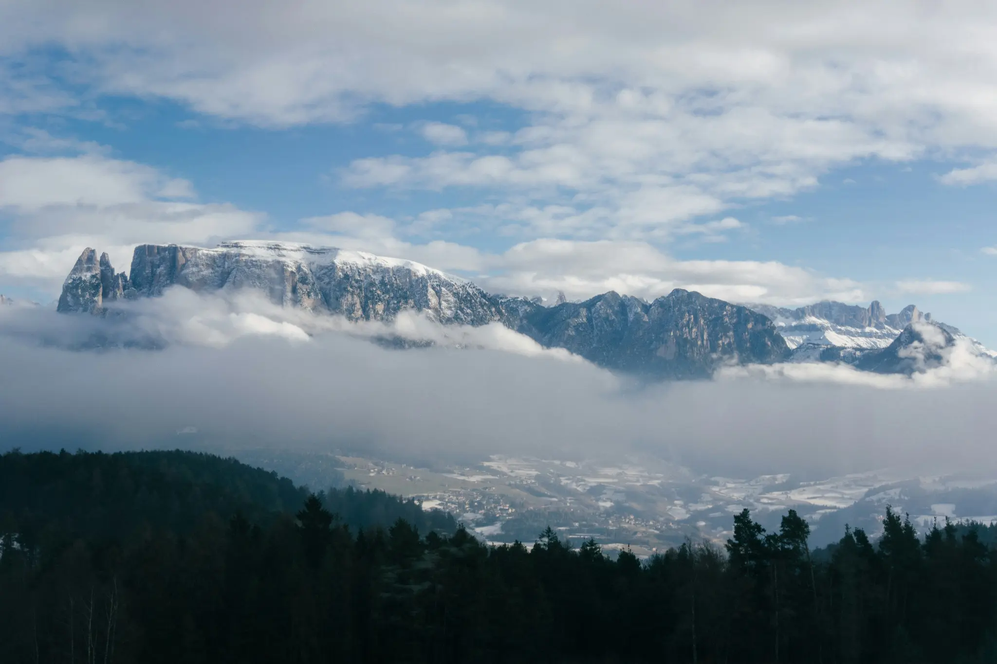 Une large photo de paysage montre des montagnes enneigées partiellement couvertes de nuages blancs, avec une forêt vert foncé