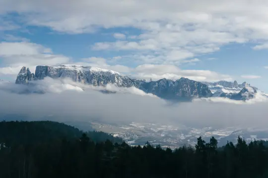 Une large photo de paysage montre des montagnes enneigées partiellement couvertes de nuages blancs, avec une forêt vert foncé