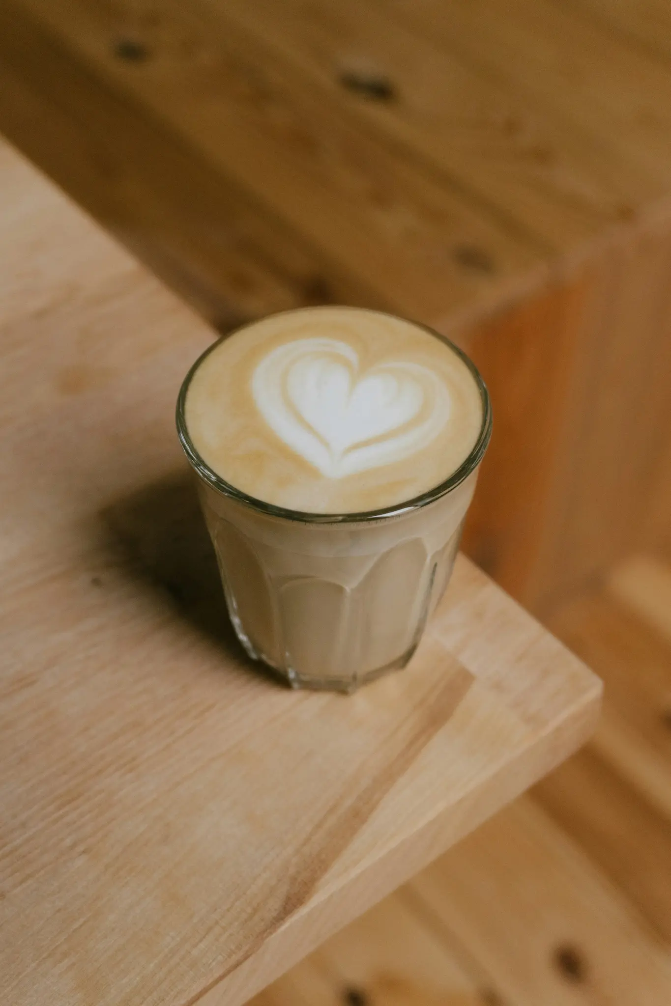 Een helder glas gevuld met een latte, met een perfect gevormd wit hart in het schuim, staat op een licht houten oppervlak.