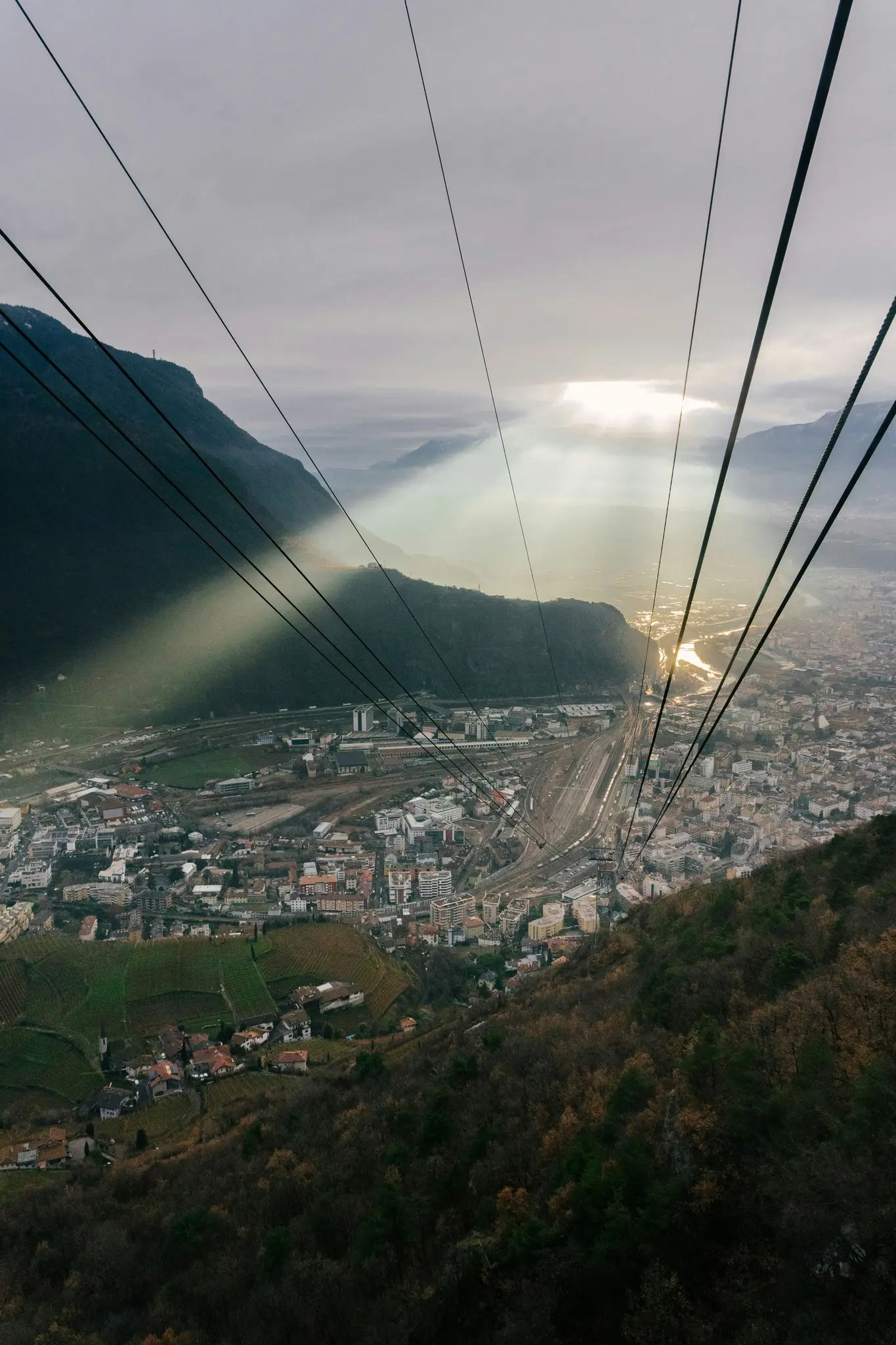 Eine Luftaufnahme von einer Seilbahn zeigt eine Stadt in einem Tal, mit Sonnenstrahlen, die durch Wolken auf die Landschaft