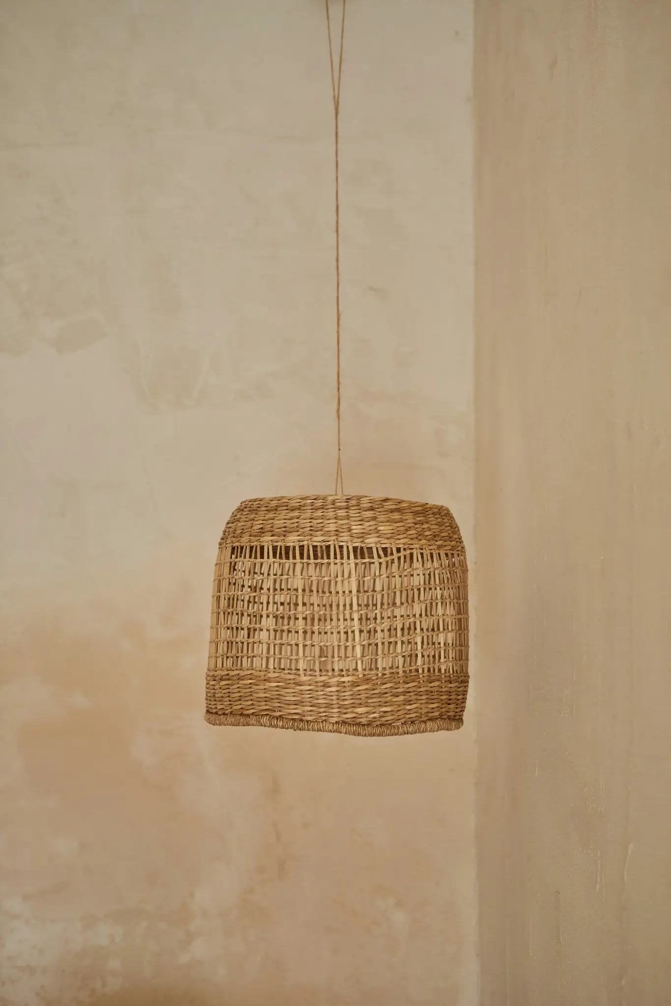Een natuurlijke, geweven rieten lampenkap hangt aan een dun touwtje tegen een getextureerde, lichtbeige muur in een hoek.