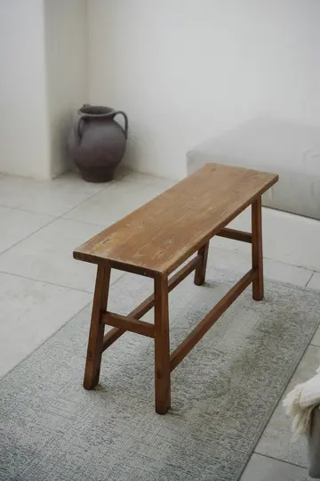 Un banco rústico de madera de diseño sencillo se asienta sobre una alfombra de color gris claro con estampado. Al fondo, una