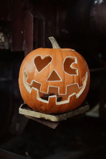 A close-up shot of a carved orange pumpkin. It has a heart-shaped left eye, a triangular right eye, and a wide, jagged…