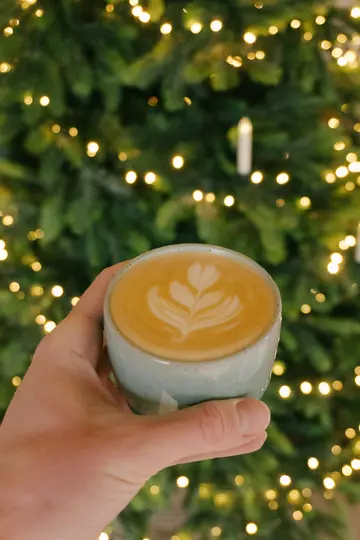A hand holds a light blue ceramic cup of latte with a heart-shaped latte art design. A blurred Christmas tree with warm…