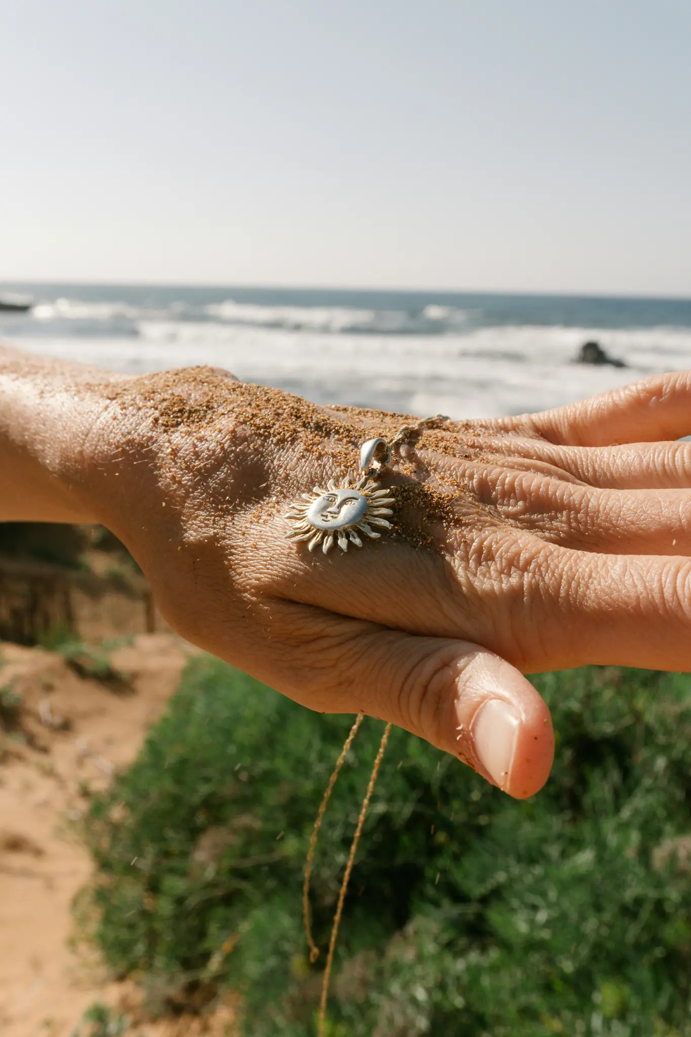 Close-up van een met zand bedekte hand die een zilverkleurige, zonvormige hanger met een lachend gezicht vasthoudt. De oceaan