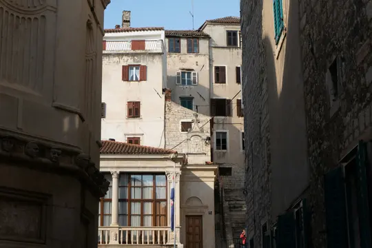 A narrow street scene showing old buildings with various windows, some with open and closed shutters, under a clear sky.