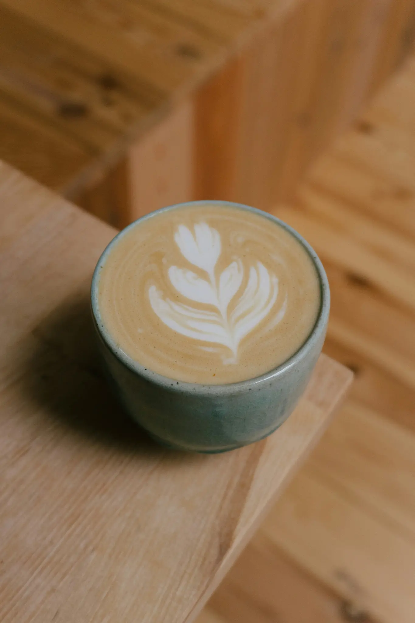 Een lichtblauwe keramische mok latte met een wit, bladvormig latte art ontwerp staat op een lichte houten ondergrond. De