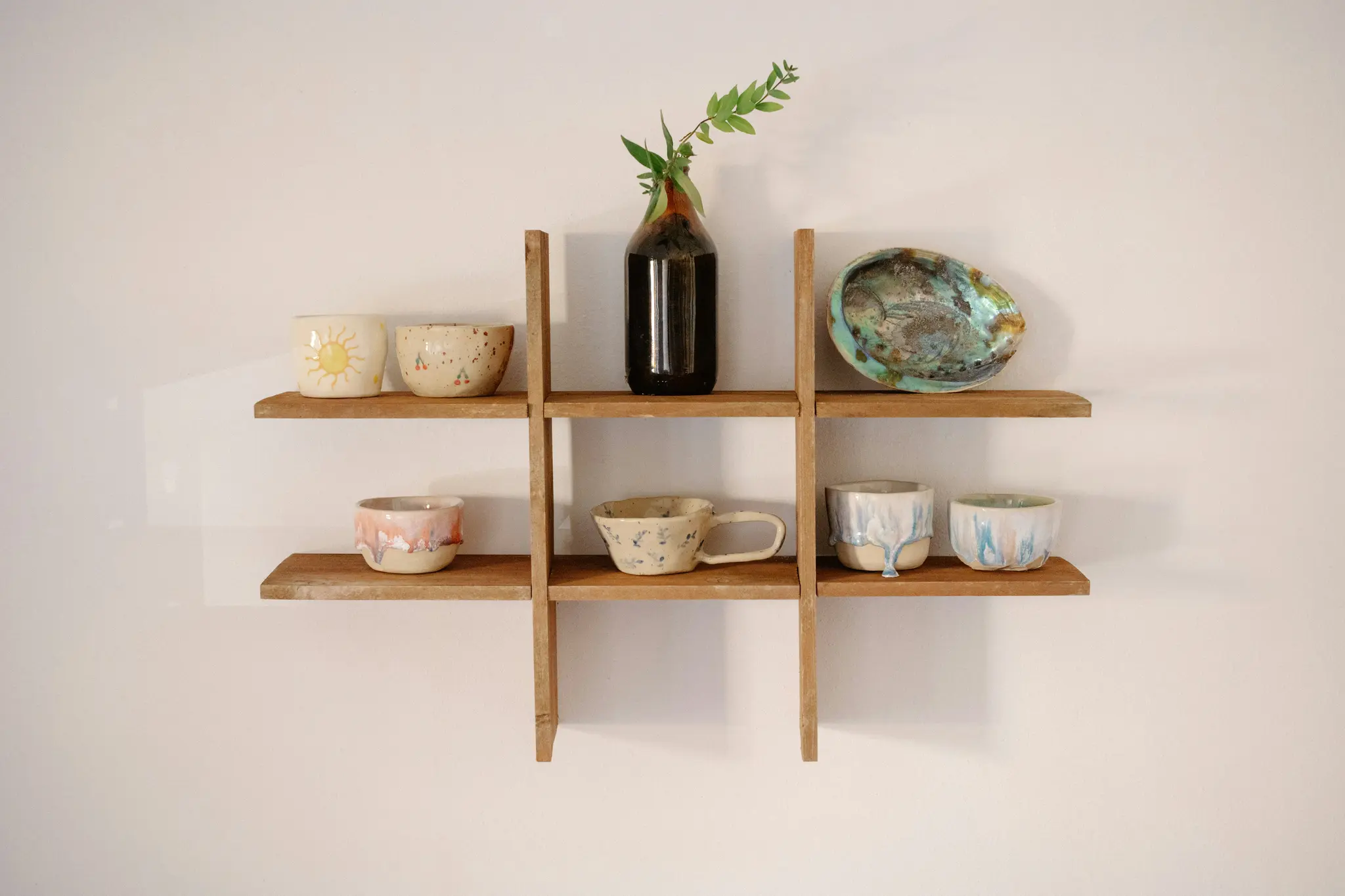 Un estante de rejilla de madera contra una pared clara sostiene varios artículos de cerámica hechos a mano, incluyendo tazas