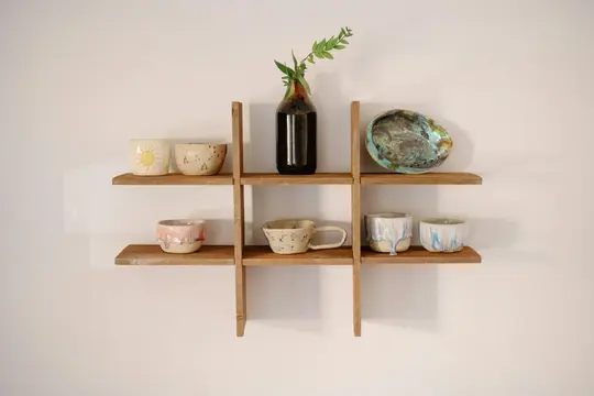 Un estante de rejilla de madera contra una pared clara sostiene varios artículos de cerámica hechos a mano, incluyendo tazas
