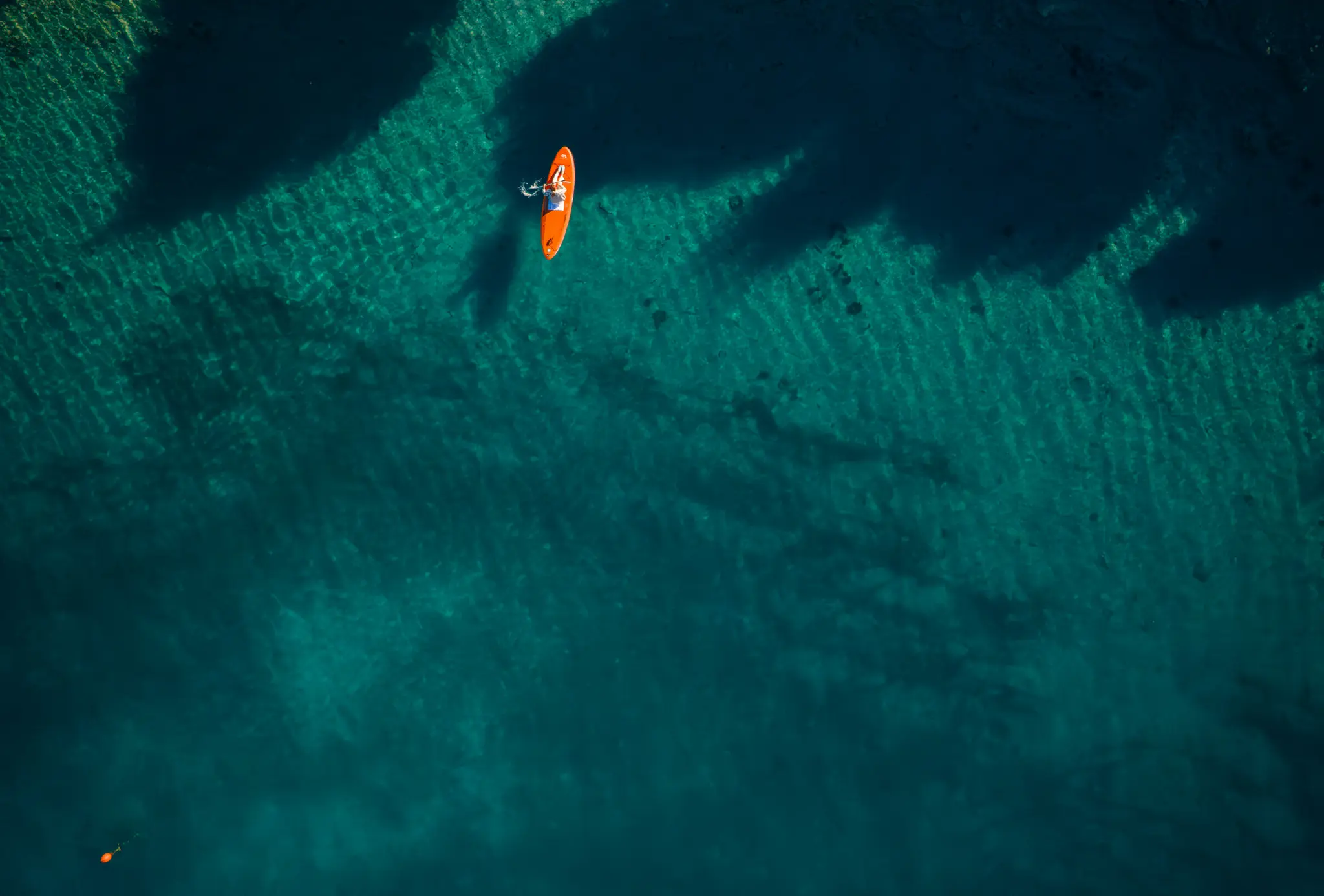 Pohled z ptačí perspektivy ukazuje osobu ležící na oranžovém paddleboardu v čisté, mělké tyrkysové vodě s tmavými stíny.