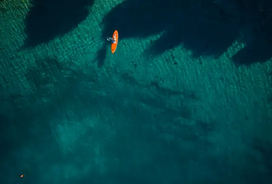 En flygvy visar en person som ligger på en orange paddleboard i klart, grunt turkost vatten med mörka skuggor.
