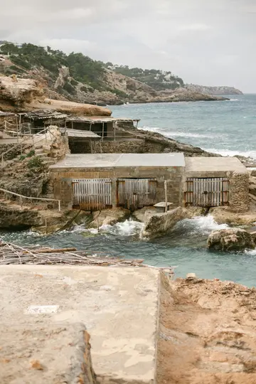 Μια θέα αρκετών ρουστίκ καλυβιών βαρκών με ξύλινες πόρτες χτισμένες σε μια βραχώδη ακτή, με τη γαλαζοπράσινη θάλασσα μπροστά.