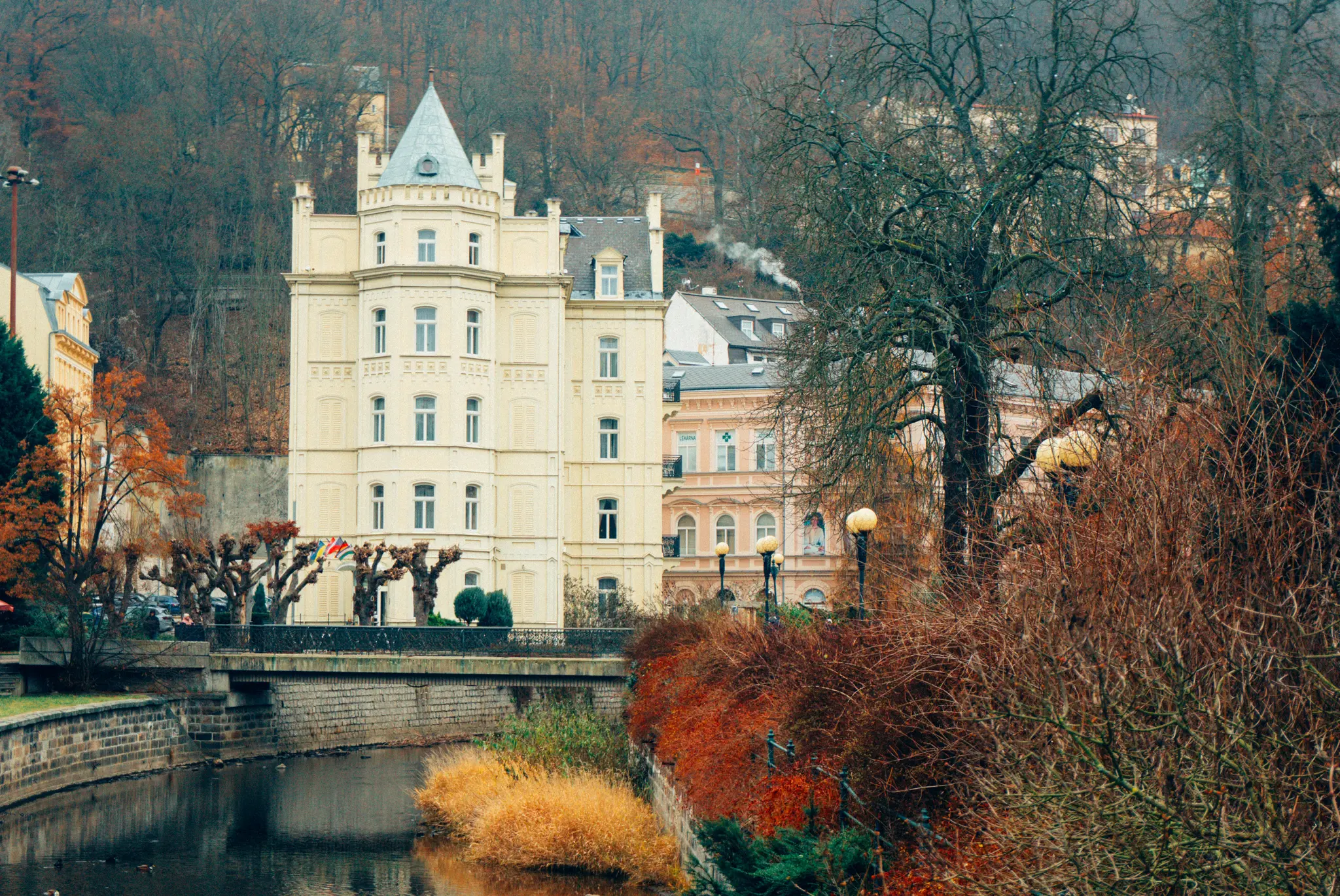 Un bâtiment de couleur claire avec une tourelle grise se dresse de manière proéminente au bord d'une rivière sombre, flanqué