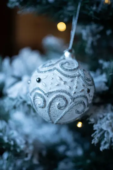 A close-up of a white Christmas ornament with silver glitter swirls and small beads, hanging on a flocked evergreen branch.