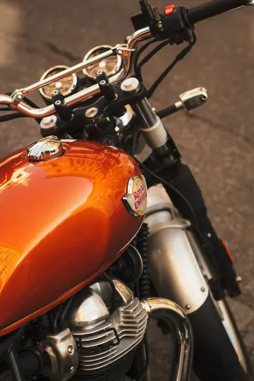 A high-angle close-up of an orange Royal Enfield motorcycle. The shiny fuel tank reflects buildings. Chrome handlebars and…